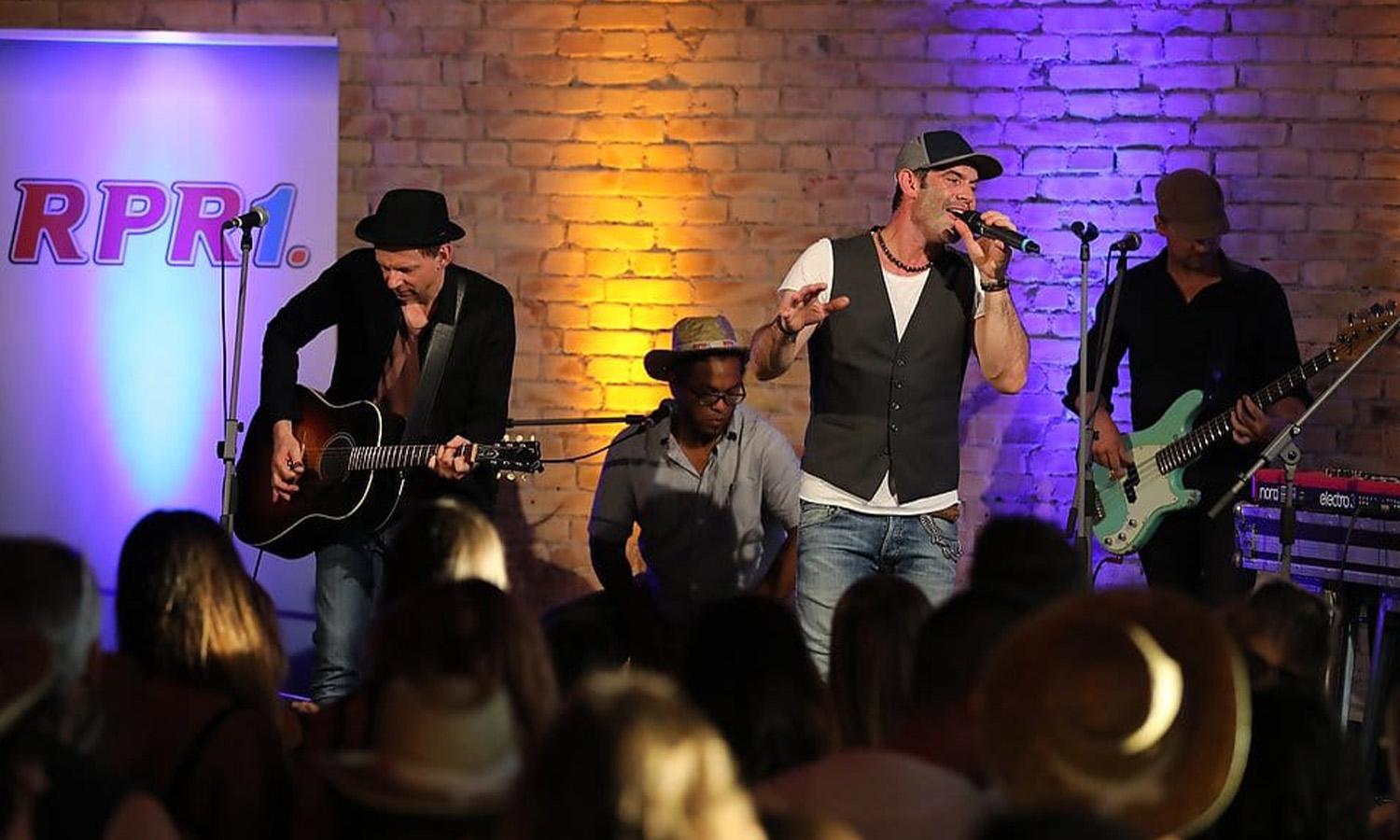 Die Band Marquess steht live auf der sommerlich beleuchteten Bühne bei RPR1. Wohnzimmerkonzert in der Manufaktur Mannheim vor tanzendem Publikum.