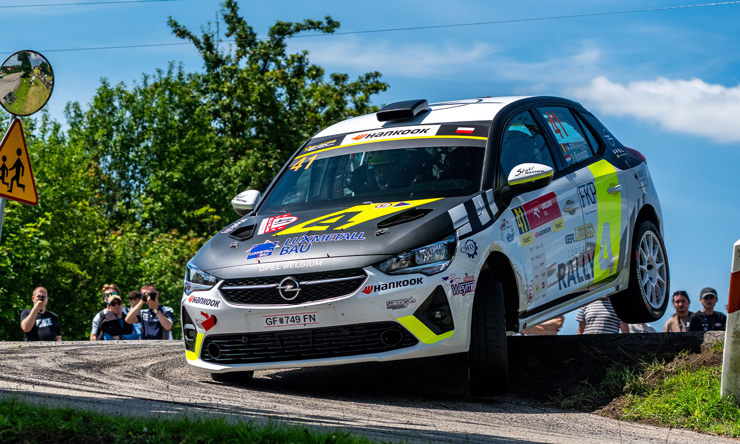 Ein Opel Corsa Rally4 driftet rasant und staubaufwirbelnd durch eine anspruchsvolle Wertungsprüfung, während das Team den Erfolg feiert.